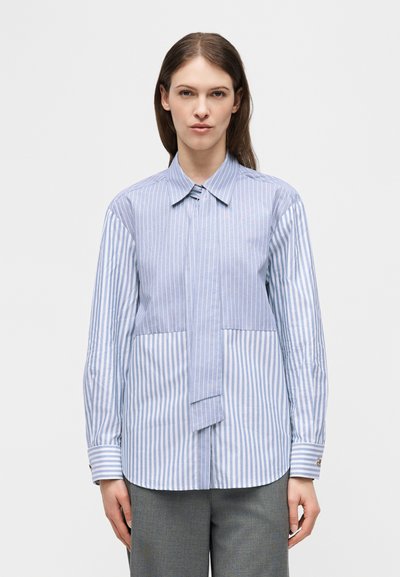 Γυναίκα με μακρυμάνικο πουκάμισο με κουμπιά, διακοσμημένο με ανάμεικτες κάθετες μπλε και λευκές ρίγες, και γκρι παντελόνι, μπροστά σε απλό φόντο.