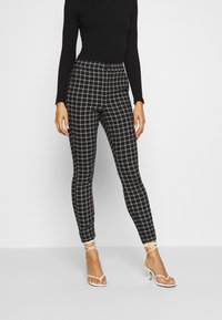 Black and gray plaid leggings with a high waist, made from ribbed fabric. Paired with white strappy high-heeled sandals.