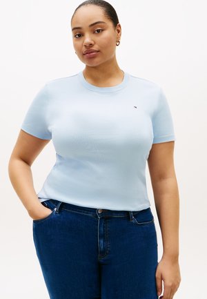 Frau mit goldenen Creolen, die ein hellblaues Kurzarmhemd und dunkelblaue Jeans trägt, steht mit einer Hand in der Tasche.