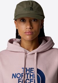 Olive green cap with a curved brim and embroidered logo. Worn with a pink hoodie featuring a blue logo. Hair styled in braids.