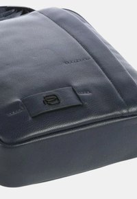 Borsa in pelle blu navy con una texture liscia, un design elegante e un accentuato logo nero sulla parte anteriore. Dettagli di cucitura sottili visibili.
