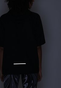 Černé sportovní tričko s krátkým rukávem a kapucí, s reflexním bílým pruhem na zadní straně. Nosené s vzorovanými legínami.