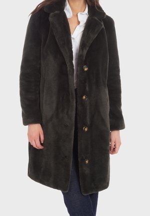 Femme portant un long manteau en fausse fourrure noire avec de grands revers, devant boutonné, chemise blanche et jean foncé sur un fond uni.