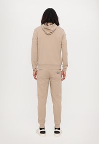 Beige mikina s kapucí a odpovídající tepláky vyrobené z měkké tkaniny, obsahující žebrované manžety a malý logo detail na zadní straně.