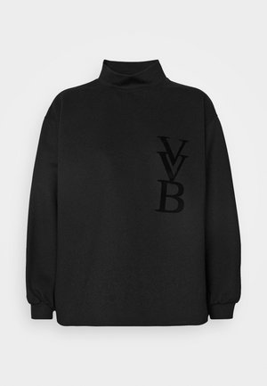 Nainen pukeutuneena mustaan collegepaitaan, jossa "LVB"-logo, valkoiset leveälahkeiset housut, mustat kärjelliset kengät, pitää käsissään beigeä takkia ja taupen väristä käsilaukkua.