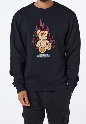 Sort sweatshirt med grafik af en bjørn omgivet af røde flammer. Detaljer inkluderer ribstrikkede manchetter og talje, med logotekst under billedet.
