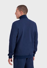 Felpa blu navy con colletto alto, polsini e orlo a costine. Texture liscia con cuciture minime. Presenta un piccolo logo vicino al colletto.