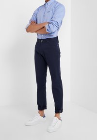 Námořnické chino kalhoty s ohrnutými lemy z bavlněné látky, doplněné světle modrou košilí s knoflíky a malým logem, a bílými teniskami.