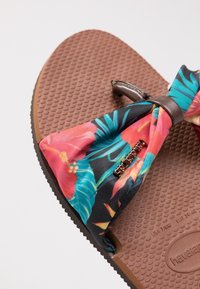 Tongs marron texturées avec un nœud floral en tissu bleu, rose et orange et un petit logo métallique de la marque sur la bride.