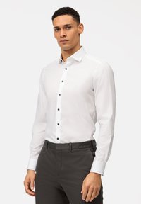 Camicia bianca con bottoni neri, caratterizzata da un colletto classico e maniche lunghe. Indossata con pantaloni grigio scuro. Tessuto di superficie liscia.
