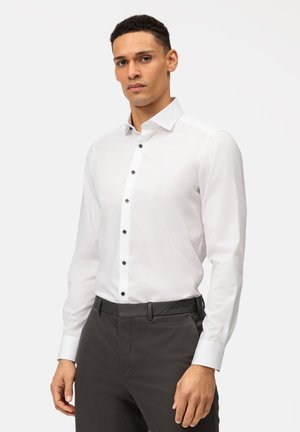 Weißes Hemd mit schwarzen Knöpfen, klassischem Kragen und langen Ärmeln. Trägt dazu eine dunkelgraue Hose. Glatte Stofftextur.