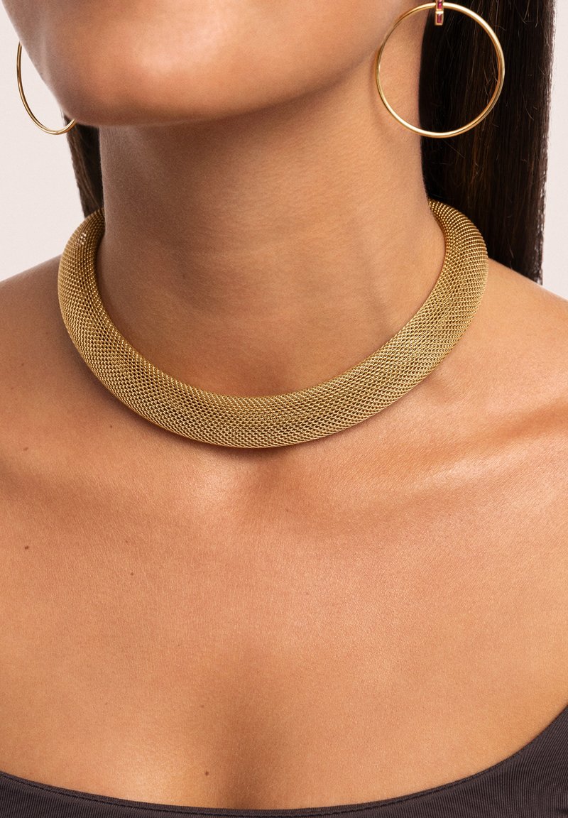 Goldenes Netz-Choker mit einem glatten, abgerundeten Design, das eine strukturierte Oberfläche zeigt. Getragen mit großen goldenen Creolen an einem nackten Hals.