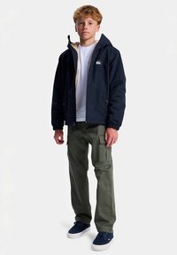 Marineblå vandtæt jakke med hætte, beige foring; hvid t-shirt under; olivengrøn cargo-bukser; marineblå sneakers med hvide såler.