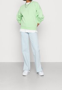 Osoba oblečená v světle zelené mikině, bílém tričku, světle modrých pruhovaných kalhotách s bočními rozparky, bílých teniskách a sytě stříbrných řetězech na bílém pozadí.