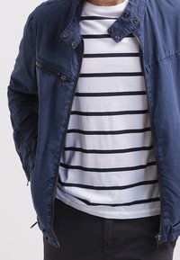 Mörkblå zip-up jacka med hög krage, med en bröstficka och en lätt, slät textur, bärs över en vit randig t-shirt.