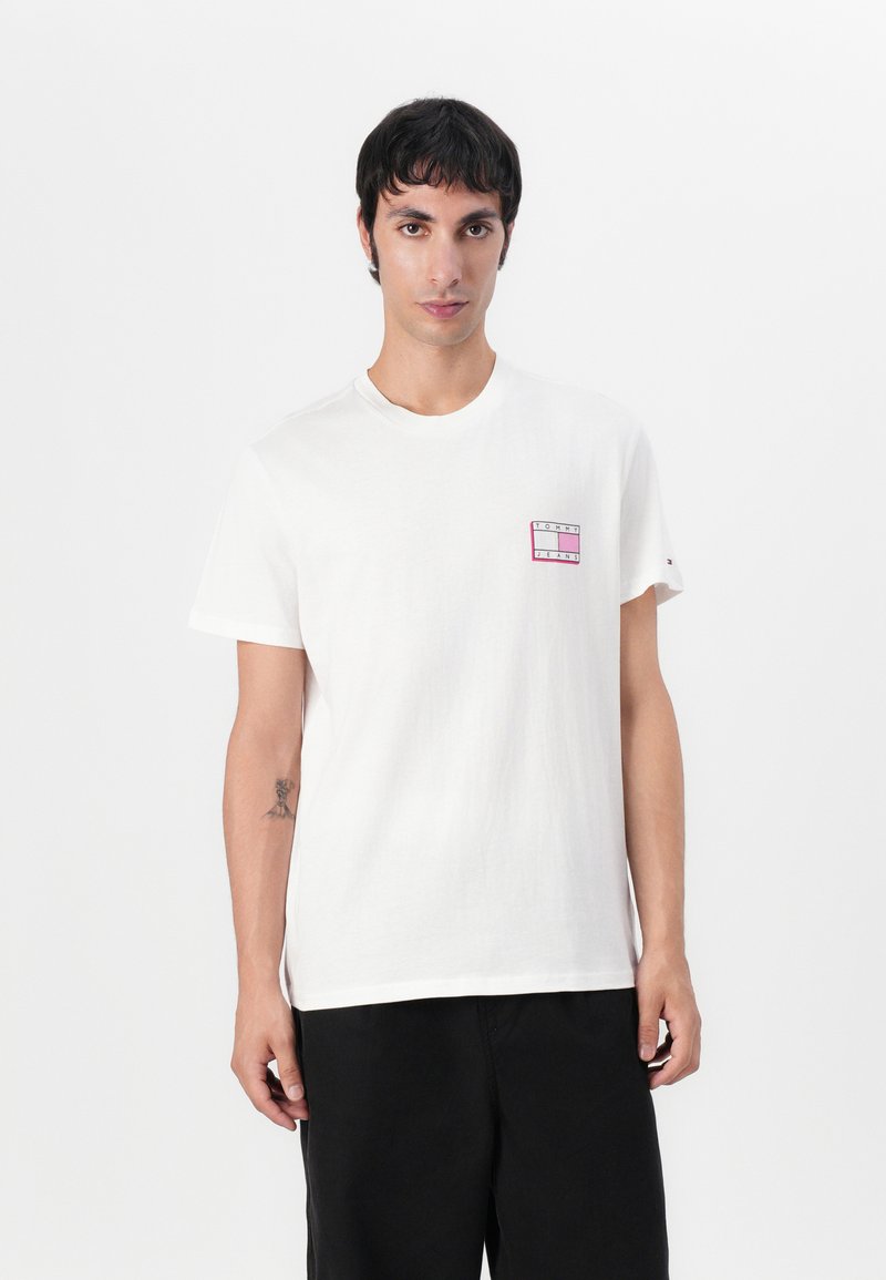 T-shirt en coton blanc avec un col rond, des manches courtes et un petit motif graphique rose sur la zone gauche de la poitrine. Pantalons noirs.