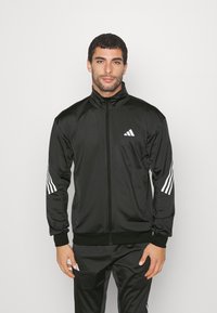 Giacca sportiva nera con colletto alto e zip completa. Presenta il logo Adidas bianco e tre strisce bianche su ciascun manica. Texture liscia.