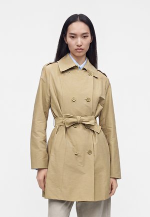 Junge Frau mit langen schwarzen Haaren, die einen beigen, doppelreihigen Trenchcoat trägt, der in der Taille gebunden ist, über hellen Hosen.