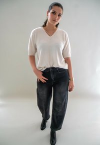 Pull en tricot blanc à manches courtes, jean large à taille haute noir et bottines noires, se tenant sur un fond clair.