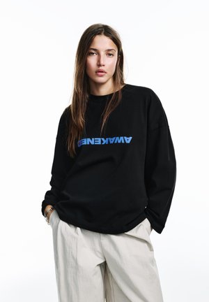 Zwarte oversized sweater met blauwe tekst waarop "AWAKENED" staat. Glad katoenen materiaal, lage schouders en wijde mouwen. Witte achtergrond.