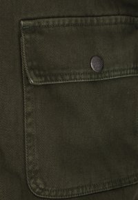 Olijfgroen stof met verticale ribbelstructuur; bevat een enkele opgestikte zak en een ronde metalen knop met reliëftekst.