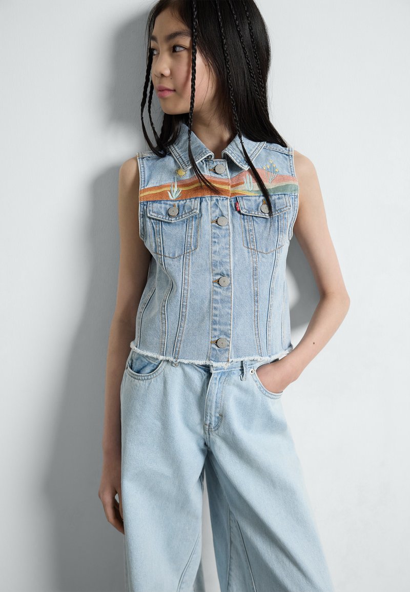 Ljusblå denimväst med ärmlös design, med flerfärgad broderad detaljering och fransade kanter, matchad med baggy jeans.