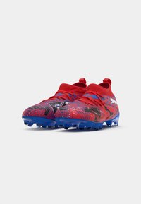 Des chaussures de football rouges avec une semelle bleue. Présentent un dessus texturé et de bold motifs noirs, avec des lacets élastiques et des languettes pour un enfilage facile.