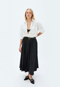 Weiße übergroße Bluse mit hochgeschlagenen Ärmeln, schwarer plissierter Maxirock, schwarzer Gürtel und spitze Flats mit Nieten. Strukturiert und fließende Stoffe.