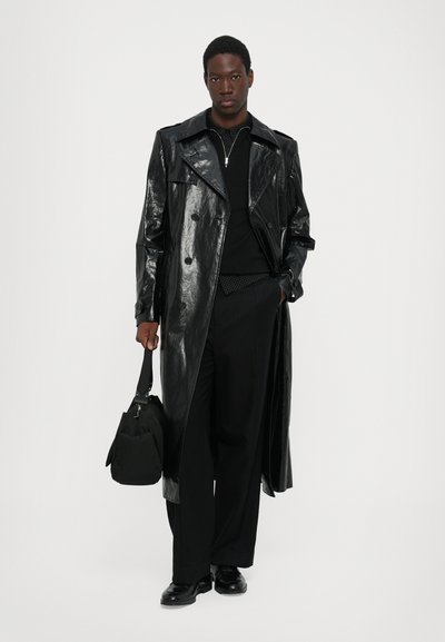 Abrigo trench de charol negro con amplios solapas y botones dobles, combinado con un cuello de tortuga negro, pantalones a medida y un bolso negro.