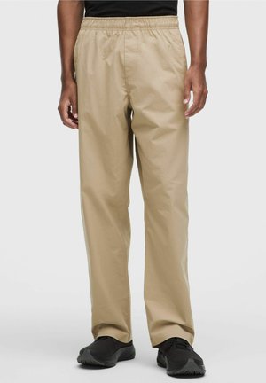 Pantalones beige con cintura elástica, usados con una camisa negra y zapatillas deportivas negras, mostrados desde la cintura hasta los pies sobre un fondo liso.