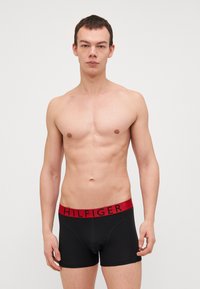 Crne bokserice s crvenim pojasom na kojem je u crnom slovu ispisano "HILFIGER". Tkanina je glatka s uskim krojem.
