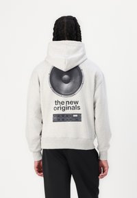 Grå hettegenser med hette, med et stort svart høyttaler grafikk og teksten "de nye originalene" på baksiden. Stoffet er mykt og strukturert.