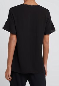 Sort bluse med korte flæseærmer, glat tekstur og dekorativ nitte-detaljering langs udskæringen. Enkel, afslappet pasform.