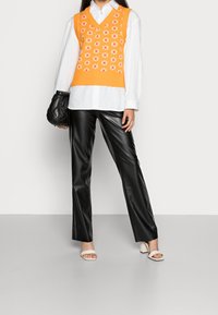 Orange stickad väst med vitt blommönster över en vit skjorta, kombinerad med svarta byxor i fejkläder och vita sandaler med blockklack.