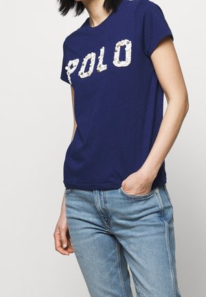 Persoon draagt een blauw T-shirt met witte letters in de vorm van schelpen die "POLO" spellen en lichtblauwe spijkerbroek, één hand in de zak, tegen een effen achtergrond.