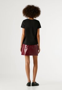 Žena s afro účesom, oblečená v čiernom top s krátkym rukávom, vínovej koženej mini sukni a čiernych mokasínach, stojí otočená chrbtom na bielom pozadí.