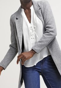 Grå skräddarsydd kappa med långa ärmar, slagkrage och rak skärning, kombinerad med en vit blus med spets och blå denimjeans.