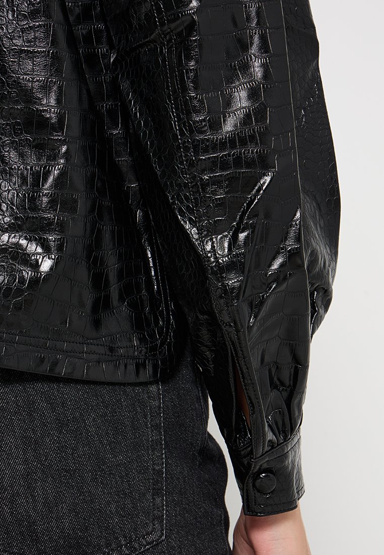 Czarna wodoodporna kurtka o fakturze krokodylej, z zapięciem na zatrzask i dopasowanym krojem. Gładkie, błyszczące wykończenie.