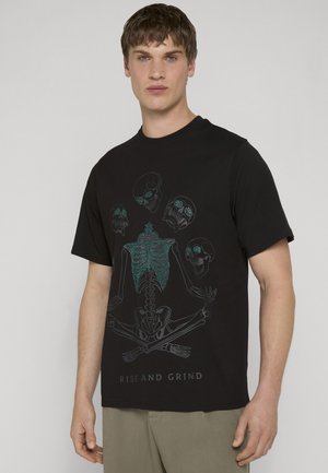 Ung mand iført sort T-shirt med et design af et skelet og kranier med grønne accenter og teksten "RISE AND GRIND."