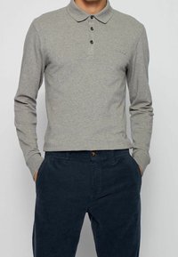 Långärmad grå pikétröja med krage, tre svarta knappar och en diskret broderad logotyp på bröstet, ihop med marinblå kordbyxor.