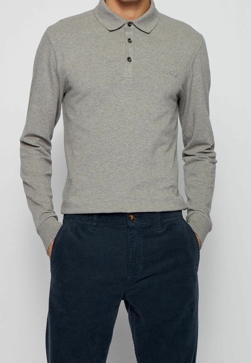 Långärmad grå pikétröja med krage, tre svarta knappar och en diskret broderad logotyp på bröstet, ihop med marinblå kordbyxor.