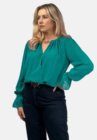 Turkusowa bluzka z dekoltem w serek, bufkami na ramionach i przezroczystymi rękawami w kształcie balonu; posiada złoty detal przy dekolcie i jest zestawiona z ciemnymi dżinsami.