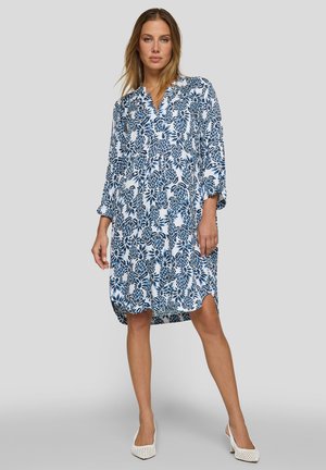 Frau trägt ein locker sitzendes weißes knielanges Kleid mit blauem Blumenmuster, dreiviertel Ärmeln und weißen perforierten Absätzen, steht vor grauem Hintergrund.