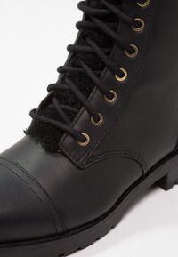 Botín negro de cuero con cordones que presenta ojales dorados, tejido negro texturizado en el tobillo y una suela de goma duradera con tracción.