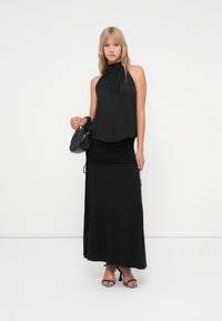 Top preto de halter com decote alto, combinado com uma saia midi preta. O modelo segura uma pequena mala preta e usa sandálias de salto pretas.