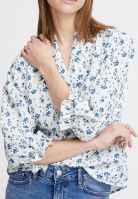 Blouse blanche avec motif floral bleu, décolleté en V et manches à volants. Associée à un jean bleu, mettant en valeur une coupe décontractée et une texture douce.