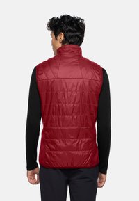 Gilet senza maniche rosso imbottito con colletto alto, caratterizzato da imbottitura trapuntata, texture liscia e tasche laterali. Indossato sopra una maglietta a maniche lunghe nera.