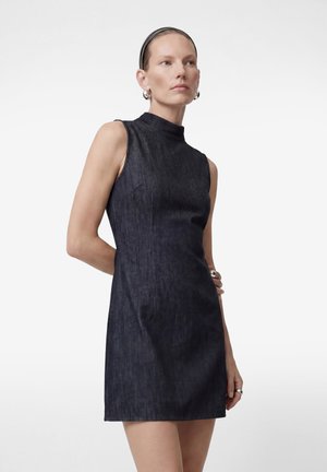 Frau trägt ärmelloses dunkles Jeans-Minikleid, silberne Creolen und ein schwarzes Stirnband, steht mit einer Hand hinter dem Rücken.
