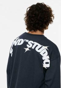 Felpa di colore blu navy scuro realizzata in cotone, con grande testo grafico bianco e motivi astratti sul retro. Vestibilità rilassata con spalle basse.