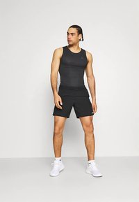 Débardeur de sport noir et short, design ajusté, texture lisse. Baskets blanches, détails de logo sur le débardeur et le short. Modèle en pose isolée.